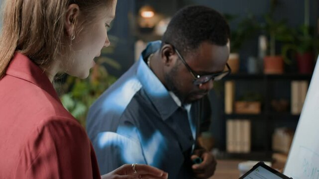 Tilt up shot of young Caucasian woman using VLM on digital tablet while talking to male African American colleague writing on flipchart in office