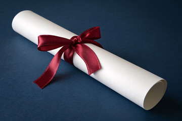 Rolled diploma tied with a red satin ribbon on dark blue background, graduation concept
