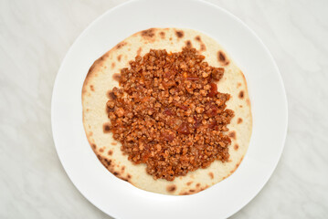 Tortilla with stewed mixed pork and beef minced meat with peeled and chopped tomatoes in tomato juice on the white plate on the light gray marble table texture. Top view.