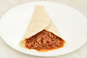 Tortilla wrap with stewed mixed pork and beef minced meat with peeled and chopped tomatoes in tomato juice on the white plate on the light gray marble table texture. Close-up. Selective focus.