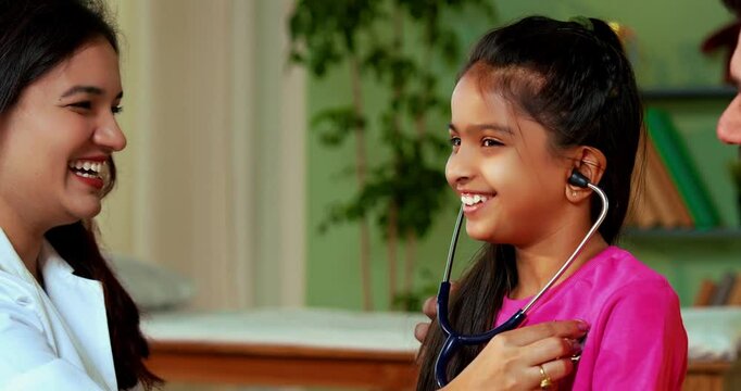 Indian Asian playful female doctor or pediatrician engaging young girl patient by letting her hear own heartbeat using stethoscope in a warm and friendly clinic atmosphere