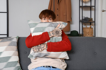 Afraid young tattooed man with pillow sitting on sofa at home