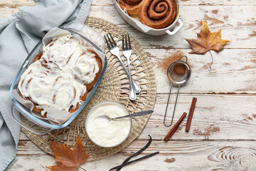 Baking dishes of tasty cinnamon rolls with cream and autumn leaves on white wooden background
