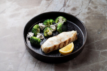 Indulge in a culinary experience featuring a tender grilled fish fillet, accompanied by vibrant broccoli and cauliflower florets topped with creamy cheese