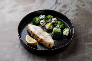 Savor a culinary masterpiece featuring tender grilled fish accompanied by bright green broccoli, elegantly presented on a dark plate. Fresh lemon adds a zesty touch to this dish