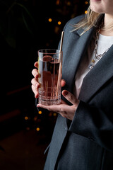 A stylishly dressed individual holds a tall glass filled with a refreshing beverage in a warmly lit European restaurant. The atmosphere is inviting, perfect for an evening out