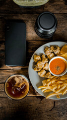 A plate of crispy side dishes featuring fried mushroom bites, French fries, and tortilla chips, served with a spicy dipping sauce. Paired with a refreshing iced drink, the meal is set on a rustic wood