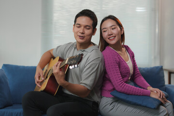 Young asian man playing acoustic guitar and singing for his girlfriend, both sitting back to back...