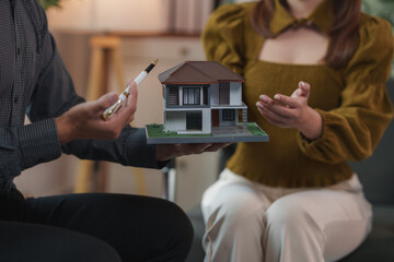 Real estate agent holding a miniature house model and a pen, presenting it to a potential female buyer who is gesturing towards the model during a meeting