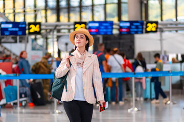 Caucasian happy young woman in straw hat holds passport with boarding pass. Defocused check flight area in background. The concept of vacation and flight trip