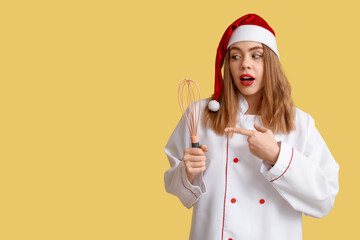 Surprised female chef in Santa hat pointing at whisk on yellow background