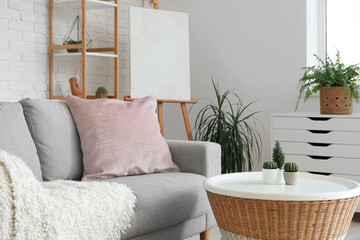 Loft style interior of light living room with cozy sofa, chest drawer, table, houseplants, shelving unit and easel
