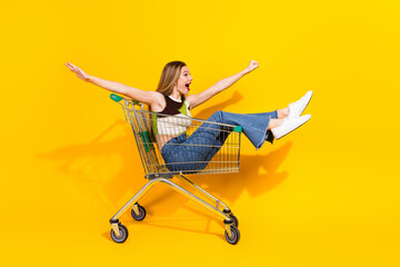 Joyful young woman riding a shopping cart on a vibrant yellow background celebrating a carefree...