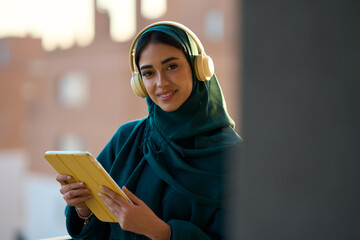 Young muslim woman wearing headphones and using tablet on the balcony