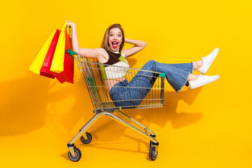 Young woman in casual style sitting in a shopping cart with paper bags against a yellow background