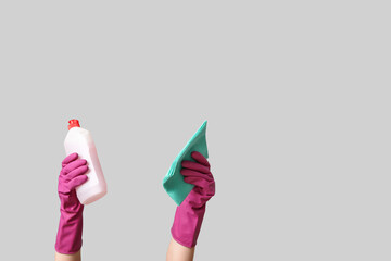 Female hands in rubber gloves with bottle of detergent and rag on grey background