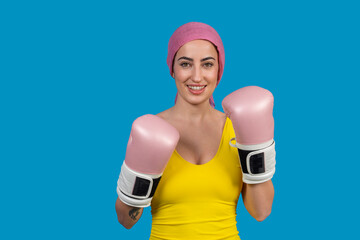 Young woman wearing pink boxing gloves and headscarf smiling on blue background fighting cancer