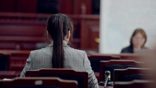 A woman sits in a courtroom with a judge and another woman. The woman in the suit is wearing a ponytail