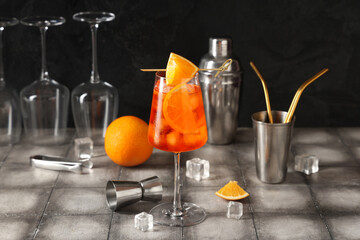 Glass of Aperol spritz cocktail with ice cubes and shaker on grey tile table