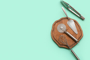 Wooden board with stainless steel pizza cutter, whisk, spatula and tongs on color background
