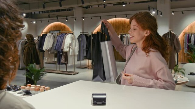 Over the shoulder view of brunette female cashier accepting non-cash payment from cheerful young woman and giving two shopping bags to her during workday
