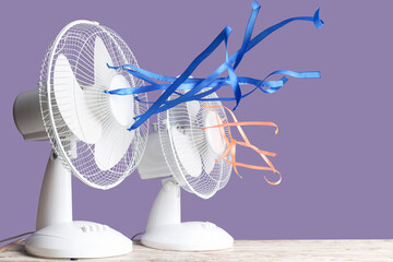 Electric fans with fluttering ribbons on table against violet background