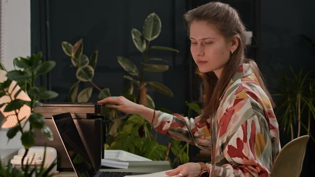 Medium shot of female Caucasian manager using printer to copy documents while sitting at desk in front of wireless laptop at office with plants