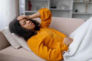Young black woman lying down on sofa with pained expression, holding her stomach and head, african american female feeling unwell at home, experiencing discomfort or illness, having acute pain