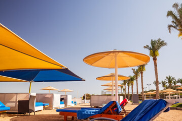 Colorful loungers and beach chairs are arranged under large umbrellas at a sunny resort beach. Blue sky and palm trees are on background. Summer vacation concept.