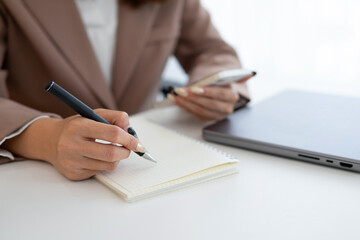 A businesswoman is sitting and recording something in a notebook. She holds a mobile phone in her hand and uses a pen to write in the notebook.