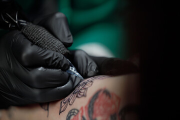 Fototapeta premium Close up tattoo master professional tattoo artist in black gloves making a rose tattoo on a man's forearm. He is holding a tattoo machine.