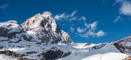 Snow covered Matterhorn mountain with ski lifts, slopes, and tracks under a vivid blue sky with scattered clouds in Cervino Ski Paradise.