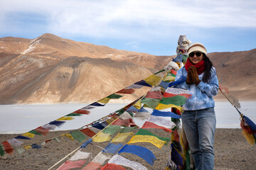 Fototapeta premium Travelers thai woman travel visit stand for take photo with Prayer flag for blessing at viewpoint with mountains and Pangong lake while winter season at Leh Ladakh in Jammu and Kashmir, India