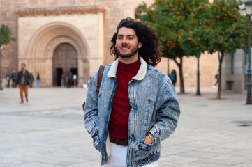 Young long-haired student walking in the city on a sunny day, wearing a denim jacket and white pants, carrying a backpack and looking away smiling