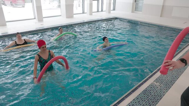 A group of women participates in an aquatic therapy session using foam noodles in a modern indoor rehabilitation pool. The session focuses on balance, strength, and low-impact resistance training in w