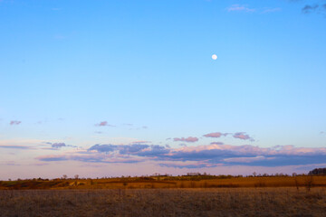 The photograph depicts a wide evening landscape with a full moon in the upper right corner of the...