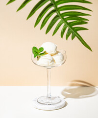 Scoops of vanilla ice cream with mint leaf in a tall glass on a beige white background with palm leaf.
