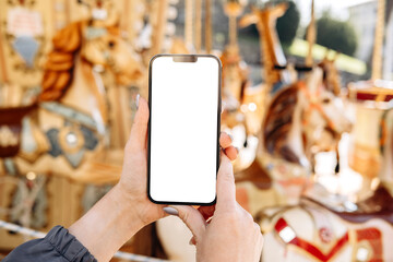 Mockup woman's hand holding using smart phone with white blank screen POV on the background of a carousel with horses, mobile app design and advertising, online marketing