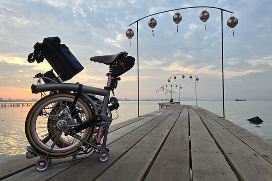 Penang, Malaysia - March 6, 2025: A serene sunrise at Lee Jetty, Penang, with a folded Brompton bicycle resting on a wooden pier.
