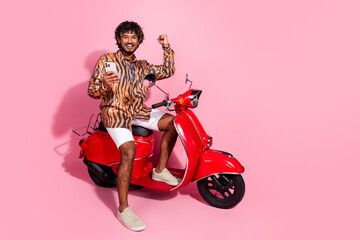 Young stylish man on pink background posing on a red scooter while holding a smartphone and showing...