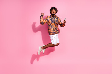 Young curly-haired man jumping against a vibrant pink wall, expressing joy and energy while holding...