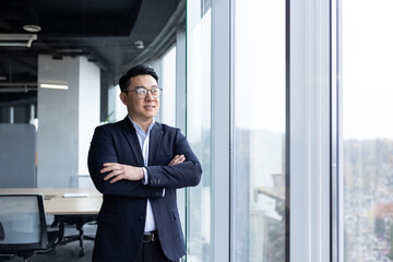 A young Asian man, founder of a company, director of a company, stands in an office in a suit, arms...