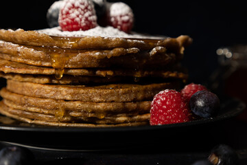 Close up of stacked pancakes with red raspberries and blueberries on top with sprinkled icing sugar on a black plate on a dark background. Homemade food, Dark food