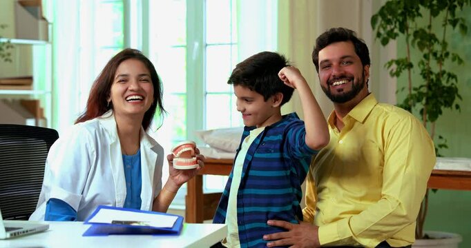 Indian Asian young female dentist demonstrating a denture model to a small boy sitting on his father&rsquo;s lap in modern clinic, as the cheerful child flexes his muscle to show strong teeth, all smiling
