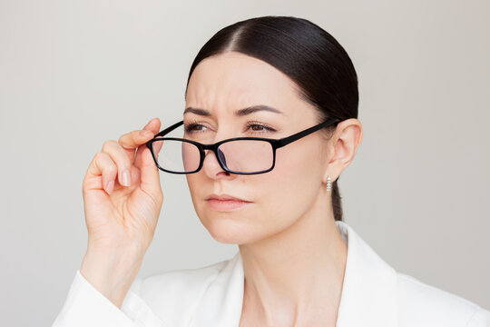 Pretty young brunette woman in white jacket experiencing vision problems, squinting while taking of her glasses