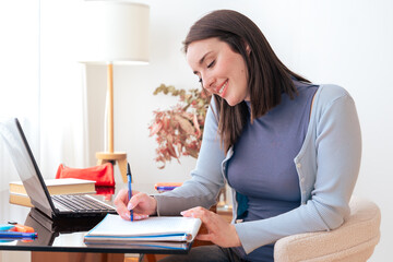 Caucasian smiling student studying at home, writing in notebook while using laptop. Modern workspace with stylish decor, lamp, and stationery. Education, learning, productivity, and concentration.
