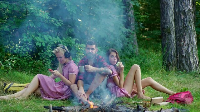 Girls and men camping. Friends enjoying a summer cam. Young friends campers sitting by the fire. Tourists in the forest. Group of friends on a camping adventure. Young People around a cozy campfire.