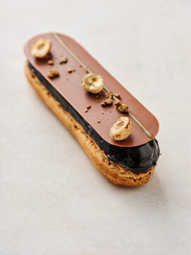 A beautifully crafted chocolate eclair features a shiny chocolate glaze topped with caramelized banana slices. The treat sits elegantly on a smooth, neutral surface, enticing dessert lovers