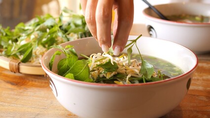 Freshly made pho soup is garnished with bean sprouts and basil leaves Woman adds ingredients that...
