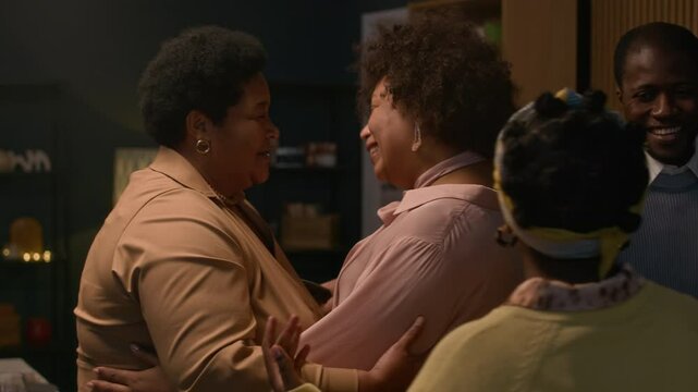 Medium shot of senior Black woman greeting adult daughter with gentle hug then siblings giving tender embrace too during festive evening dinner at home, heartfelt family moment